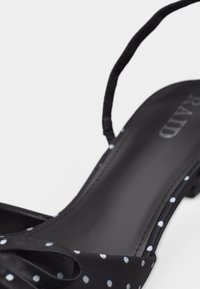 Μαύρο πέδιλο με ανοιχτά δάχτυλα, λευκές πουά, χαμηλό τακούνι και λουράκι στον αστράγαλο, με έμφαση στη σόλα και στη λεπτομέρεια στο πλάι.