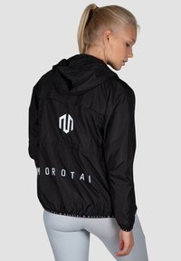 Sort vindjakke med hætte, med et logo på ryggen. Letvægtsmateriale med elastiske manchetter og talje, glat tekstur.