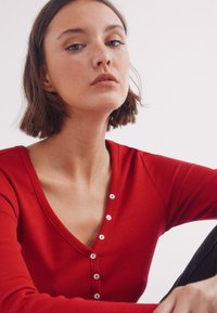 Rode geribbelde longsleeve top met een diepe V-hals en vijf witte knopen aan de voorkant. Glad textuur en een aansluitend ontwerp.