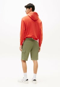 Sudadera roja con capucha, pantalones cortos de carga oliva con bolsillo lateral, calcetines blancos y zapatillas negras y blancas. Fondo neutro.