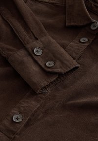 Brązowa koszula sztruksowa z kołnierzykiem i mankietami na guziki, czarne guziki i teksturowany wzór tkaniny.