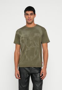 Πράσινο χακί T-shirt με κοντά μανίκια, με διακριτικό μοτίβο και μικρό λογότυπο "DIESEL" στο στήθος. Συνδυάζεται με μαύρο δερμάτινο παντελόνι cargo.
