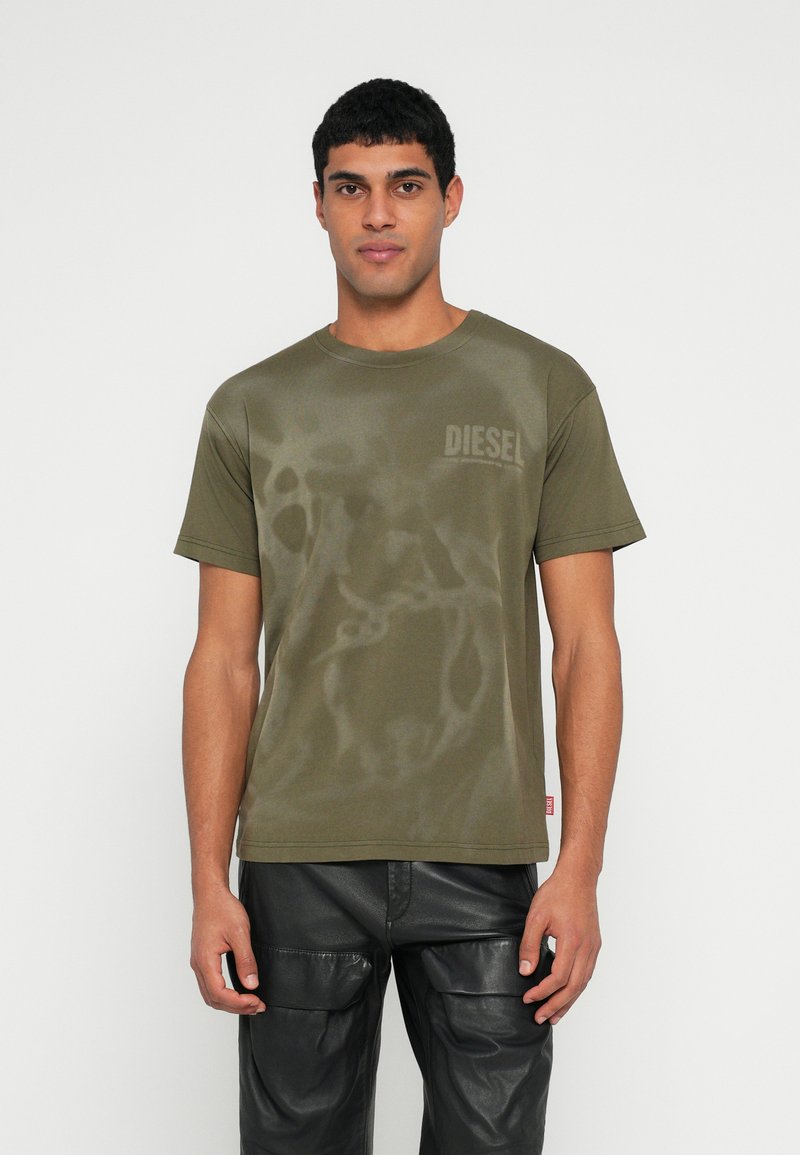 Πράσινο χακί T-shirt με κοντά μανίκια, με διακριτικό μοτίβο και μικρό λογότυπο "DIESEL" στο στήθος. Συνδυάζεται με μαύρο δερμάτινο παντελόνι cargo.