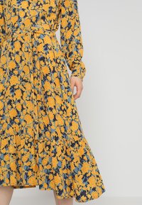 Vestido floral con patrones en amarillo y azul sobre un fondo marino, de manga larga, con cintura ajustada y dobladillo con volantes.