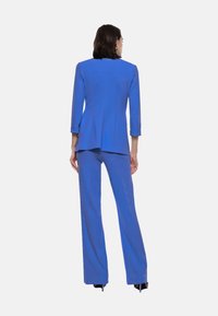 Completo blu su misura con giacca aderente e pantaloni a gamba larga, realizzato in tessuto morbido. Presenta maniche a tre quarti e dettagli minimali.
