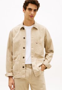 Beige Jeansjacke mit Knöpfen, zwei Vordertaschen und einem kleinen Logo. Wird über einem weißen T-Shirt getragen und zeichnet sich durch ein schlichtes, strukturiertes Design aus.