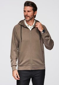 Sudadera con cremallera color beige, con capucha ajustable, bolsillos laterales con cremallera y puños acanalados. Fabricada en tejido suave, presenta detalles del logo en la manga.