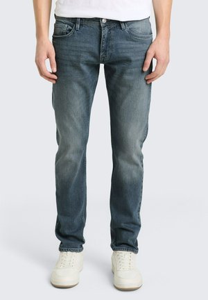 Mężczyzna w wyblakłych niebieskich obcisłych jeansach i białych trampkach, stojący na tle jednolitego jasnego tła.