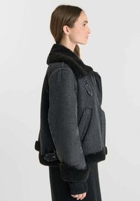 Giacca di jeans nera con colletto e polsini in pelliccia nera, dotata di tasche laterali e silhouette ampia. Texture morbida e hardware resistente in evidenza.