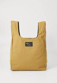 Sac en toile kaki avec un grand dessus ouvert, de forme carrée, et une étiquette noire à l'avant. Texture lisse avec des coutures renforcées.