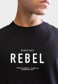 Crna pamučna majica s okruglim izrezom, s istaknutim bijelim natpisom: "REDEFINE REBEL" i geografskim koordinatama. Standardni kroj.