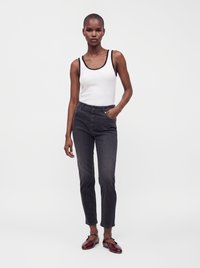 Femme portant un débardeur blanc avec une bordure noire, un jean taille haute foncé et des chaussures Mary Jane bordeaux, debout avec une main dans la poche.