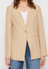 Kvinna iklädd beige randig kavaj över vit v-ringad t-shirt och blå jeans, stående med armarna avspänt längs sidorna.