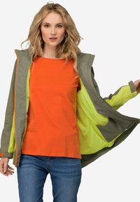 Chaqueta resistente al agua en verde olivo con forro amarillo neón. Presenta una camiseta de manga corta naranja y jeans azules. Diseño ligero.