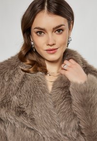 Faux fur frakke i lys brun med en blød tekstur. Har en dyb V-halsudskæring og bred krave. Modellen bærer lagdelte sølvhalssmykker og hoop øreringe.