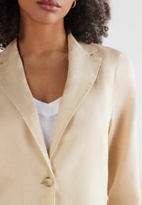 Beige linnen blazer met notch-revers, enkele knoopsluiting en gestructureerde stof. Gelaagd over een witte V-hals top.