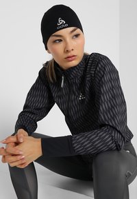 Zwarte sportjack met een hoge kraag, voorzien van een geometrisch patroon in grijstinten. Het model draagt een bijpassende zwarte beanie.