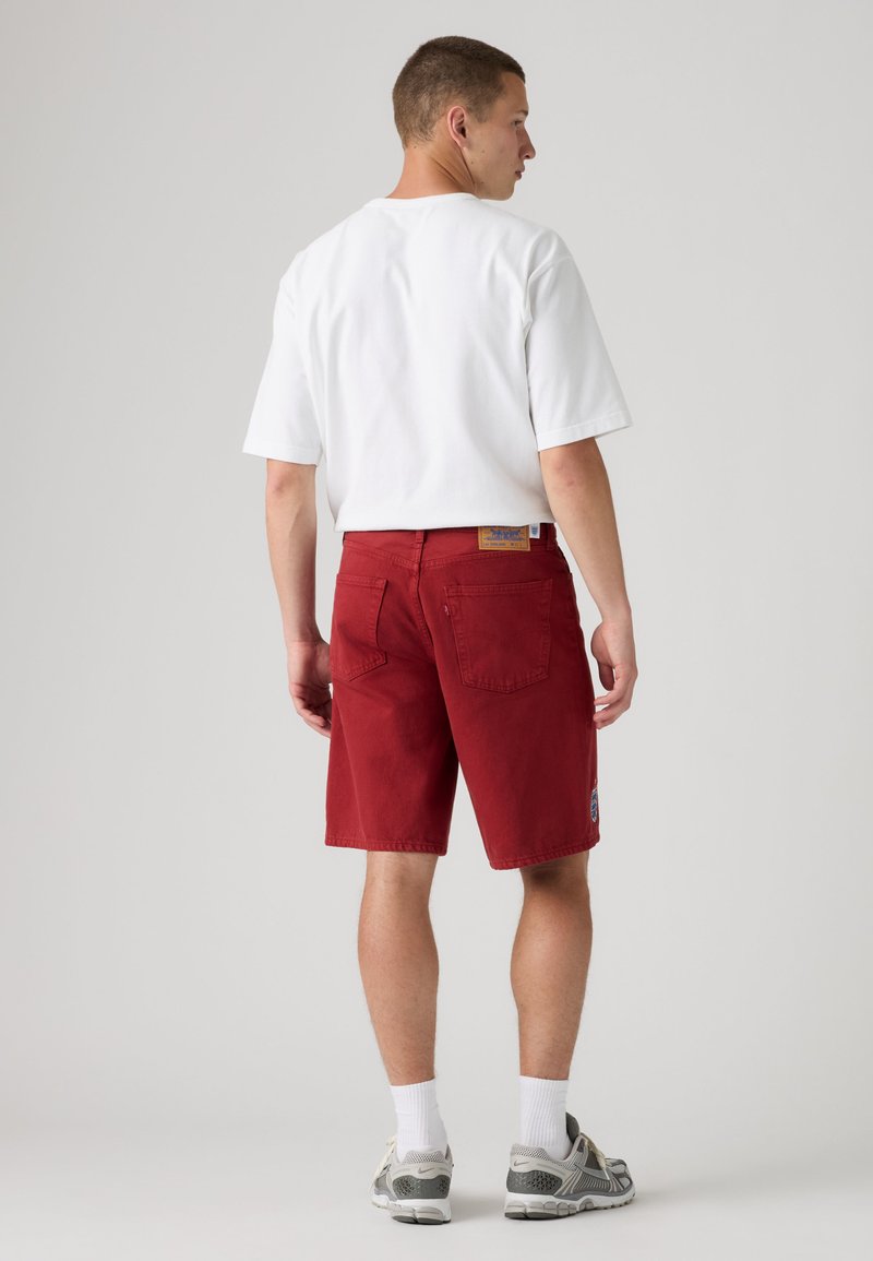Hombre vistiendo camiseta blanca, pantalones cortos rojos hasta la rodilla con bolsillos traseros, calcetines blancos y zapatillas grises, de pie de espaldas.