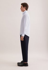 Hellblaues, gemustertes Hemd mit Knöpfen, dunkle Hose und schwarze Schuhe. Das Hemd hat einen Kragen und sichtbare seitliche Schlitze, taillierte Passform.