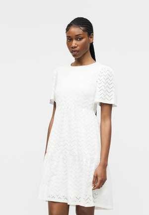 Kobieta z warkoczami, ubrana w białą sukienkę do kolan z krótkimi rękawami i wzorem otworów w kształcie zygzaka na jednolitym tle.