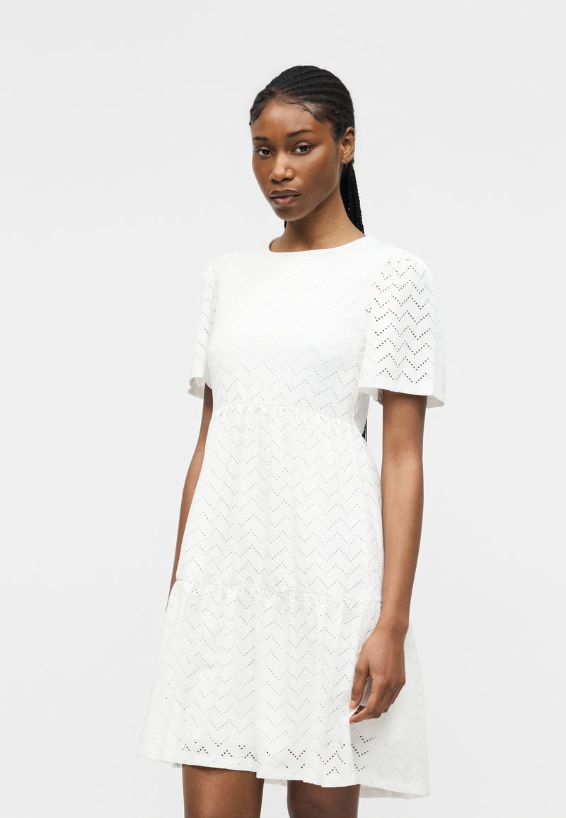 Γυναίκα με κοτσίδες που φοράει λευκό φόρεμα έως το γόνατο με κοντά μανίκια και ζιγκ-ζαγκ σχέδιο με τρύπες σε μονόχρωμο φόντο.