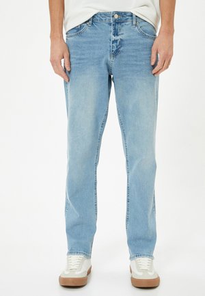 Jeans de mezclilla azul claro con corte recto, con un ligero desgaste, cierre de botón y cinco bolsillos, combinados con zapatillas blancas y marrones.