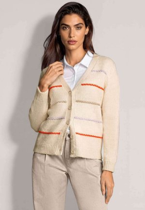 Frau trägt eine beige geknöpfte Strickjacke mit horizontalen orangefarbenen und violetten Streifen über einem weißen Hemd mit Kragen und hellbeigen Hosen.