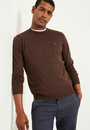 Brauner Pullover mit Rundhalsausschnitt, gerippten Bündchen und Saum, getragen über einem weißen Hemd. Das Material wirkt glatt und gestrickt.