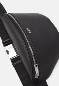 Borsa a tracolla nera in pelle con superficie testurizzata, zip argentata e tracolla nera regolabile con logo in rilievo. Forma liscia e curvilinea.