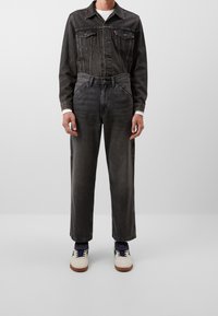 Chaqueta de mezclilla gris oscuro con dos bolsillos en el pecho, combinada con jeans gris oscuro ajustados. Camisa blanca por debajo y zapatillas crema con detalles azules.