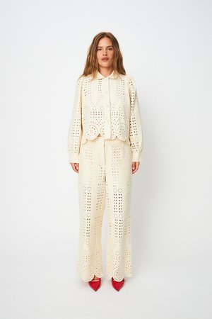 Naine kannab kreemvalget nööbitavat pitspluusi ja sobivaid laia säärega pükse lainekujuliselt lõigatud äärega, punaste teravate kingadega, seisab sirgelt.