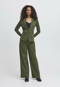 Completo su misura verde oliva con giacca a un bottone e pantaloni a gamba larga. Tessuto morbido, design minimalista, nessun motivo o accento visibile.