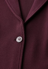 Detailní záběr na texturovanou fialovou pletenou bundu s bílým knoflíkem označeným "street One", který zapíná přední část.