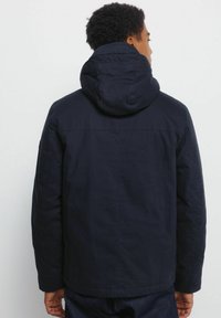 Uomo di spalle che indossa una giacca a cappuccio blu scuro con maniche lunghe, su uno sfondo chiaro e neutro.