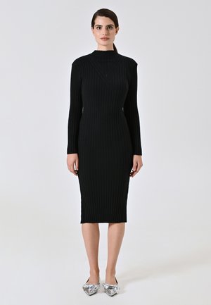Donna in piedi, che indossa un abito midi a maniche lunghe nero aderente con scollo alto e ballerine argentate con fiocchi.
