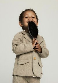 Bambino con una giacca di suit marrone chiaro a due bottoni, che tiene in mano una spazzola per capelli nera con manico testurizzato. Sfondo neutro.