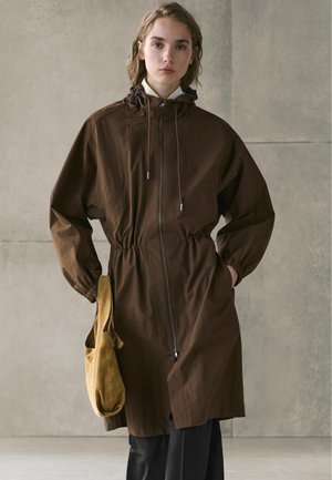 Młoda osoba w brązowym płaszczu z zamkiem i kapturem, z wcięciem w talii, trzymająca jasnobrązową torebkę na ramię, stojąca na tle gładkiej betonowej ściany.