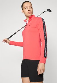 Lehká korálová zipová bunda s dlouhými rukávy a logem na bocích, v kombinaci s černými šortkami; drží golfovou hůl.