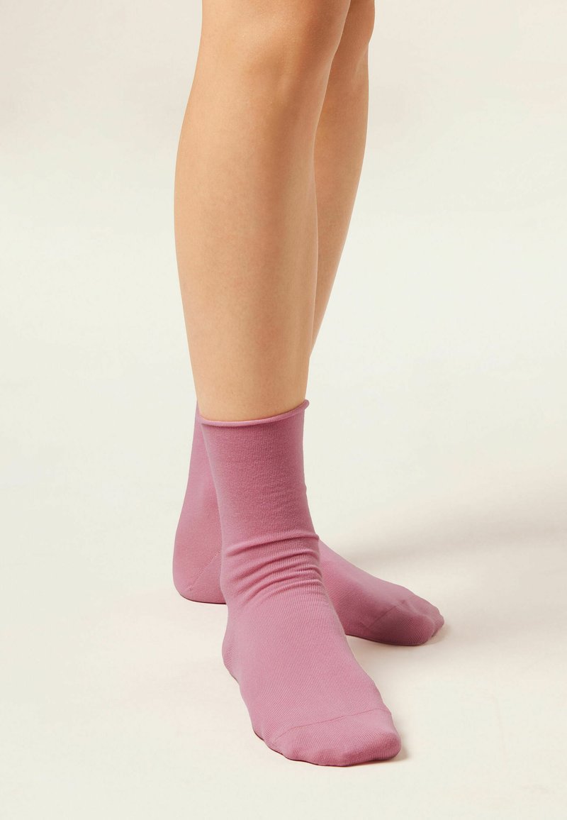 Jambes portant des chaussettes roses mi-mollet, debout sur un fond clair et neutre, montrant de légers plis sur le tissu.