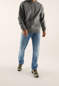 Felpa grigia in cotone con tasca frontale, jeans in denim blu e sneakers verde mimetico con dettagli neri, il tutto su uno sfondo neutro.