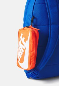 Un sac à dos bleu en tissu matelassé avec des bretelles ajustables, doté d'une pochette zippée orange attachée affichant un logo Nike blanc.