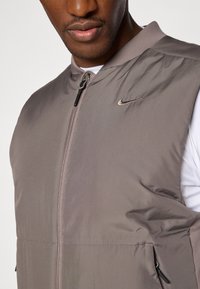Gilet senza maniche Nike, leggero, grigio con una texture liscia. Presenta tasche con zip e un piccolo logo sul petto.