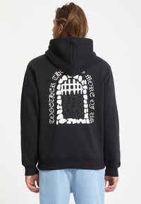 Sweat-shirt à capuche noir avec un grand graphisme blanc représentant une arche en pierre avec une toile d'araignée et du texte au dos. Matière en coton.