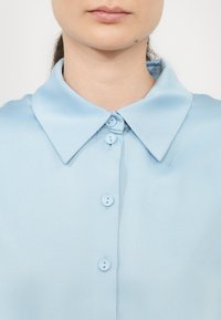 Világoskék blúz klasszikus gallérral, gombos elülső résszel és sima, texturált anyaggal. Részletei között található három gomb és rendezett kivitel.