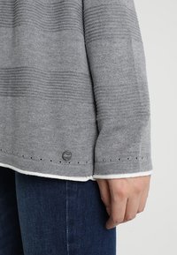 Grå stickad tröja med horisontella ränder, vit kant och liten logotypetikett. Har delikata perforeringar nära fållen och ärmslutet.