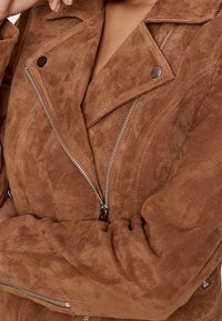 Veste de moto en daim marron avec fermeture à glissière asymétrique, revers angulaires et accents en métal argenté. Texture douce sur l'ensemble.