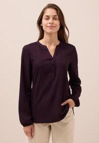 Eine Frau trägt eine dunkelviolette Bluse mit langen Ärmeln und Knöpfen sowie beige Hosen. Sie steht mit einer Hand in der Tasche vor einem schlichten Hintergrund.