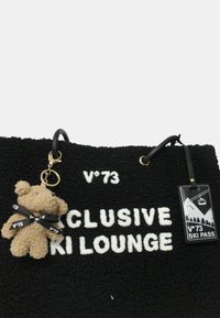 Borsa nera testurizzata con la scritta "V°73 Exclusive Ski Lounge", un piccolo portachiavi a forma di orsetto e un'etichetta del pass sciistico attaccata ai manici.