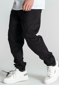 Pantalones de mezclilla negros con un corte relajado, que presentan una textura suave y detalles de costura sutiles. Combinados con zapatillas blancas marcadas "GIANNI."