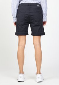 Tmavě modré kraťasy s ohrnutými lemy a zadními kapsami, vyrobené z bavlněné látky. Nošeny s bílými sportovními botami, viditelnými zezadu.